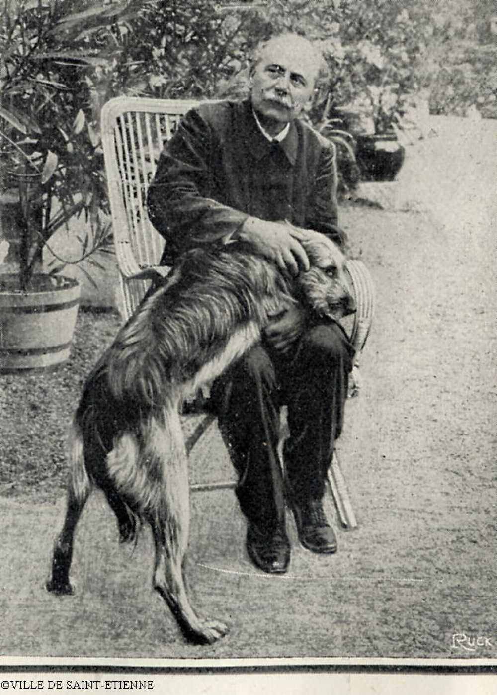Jules Massenet assis sur une chaise en osier caressant un chien lui montant sur les genoux.<br>
